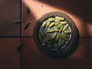 a decorative plate filled with green cardamom pods. The plate, adorned with intricate patterns in red, green, and white, sits on a tiled surface. The lighting creates a dramatic effect, with shadows casting on part of the plate and the floor.